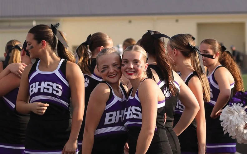 A group of adults wearing matching purple and black cheerleading uniforms with BHSS on the front are standing together outdoors, some smiling and posing for the camera. Description generated by AI