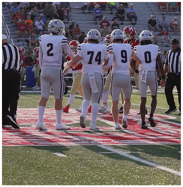 A group of people in football uniforms and a referee stand on a football field during a game with spectators in the background. Description generated by AI