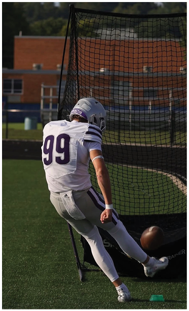 A person wearing a white football jersey with the number 99 and a silver helmet is standing next to a net on a sports field. Description generated by AI