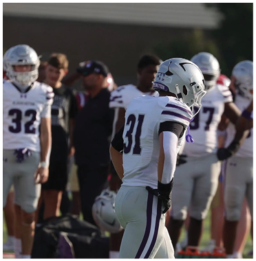 A group of people on a sports field with one person in a white football uniform and helmet numbered 31 standing in the foreground. Description generated by AI