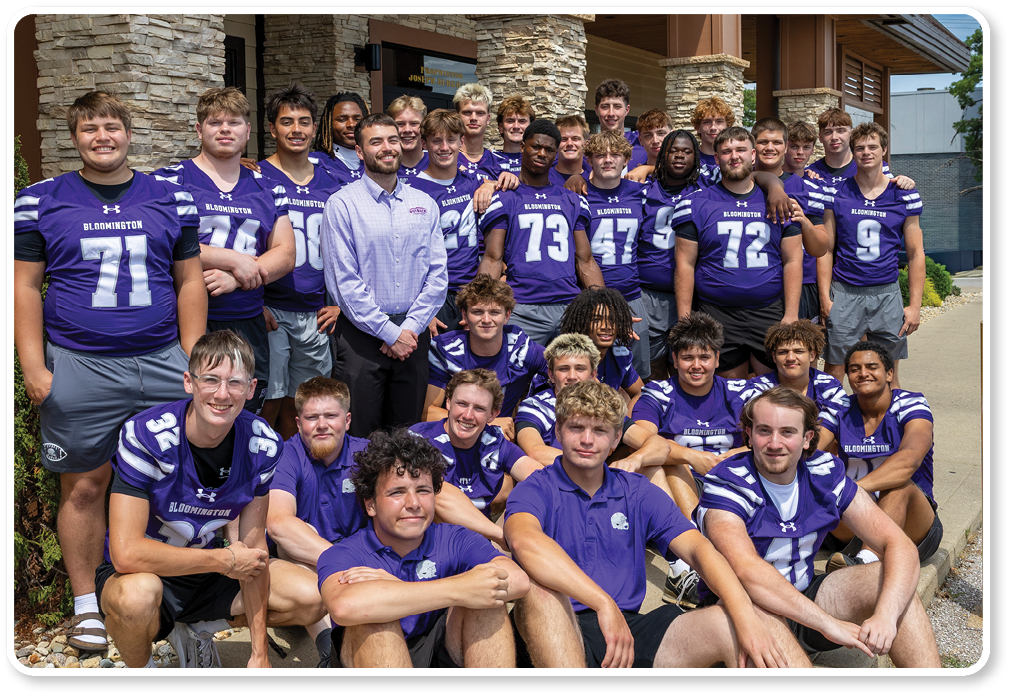 A group of people wearing matching purple sports jerseys and one person in a light purple shirt pose together outside an Outback Steakhouse building on a sunny day. Description generated by AI