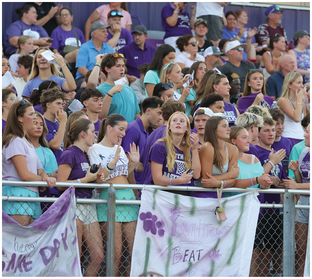 A crowd of people wearing purple and teal shirts are gathered behind a fence with hand-painted banners showing support for a sports event. Description generated by AI