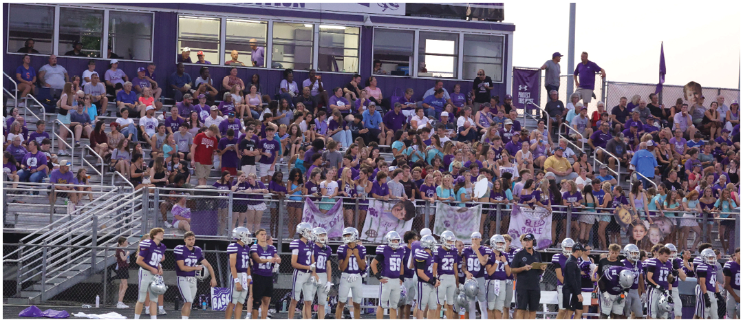 A large crowd of people wearing purple and white is gathered in bleachers at a football stadium under a banner that reads “Panther Football Tradition." Description generated by AI
