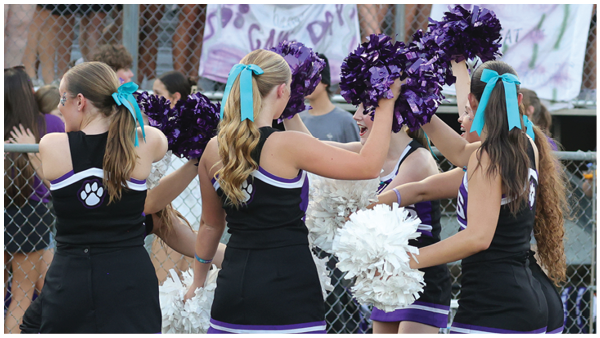 Four people with long hair tied with blue ribbons are dressed in black cheerleading uniforms and holding purple and white pom-poms while performing at an outdoor event with a chain-link fence and spectators in the background. Description generated by AI