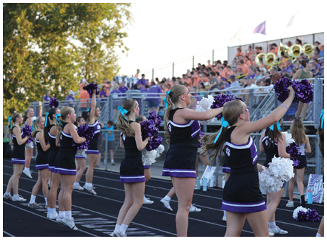 A group of adults in matching cheerleading uniforms with blue hair ribbons perform a routine on a track in front of a crowded stadium. Description generated by AI