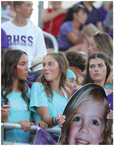 A group of people wearing casual clothing are gathered closely together at an outdoor event, with some holding a large cutout of a smiling child's face. Description generated by AI