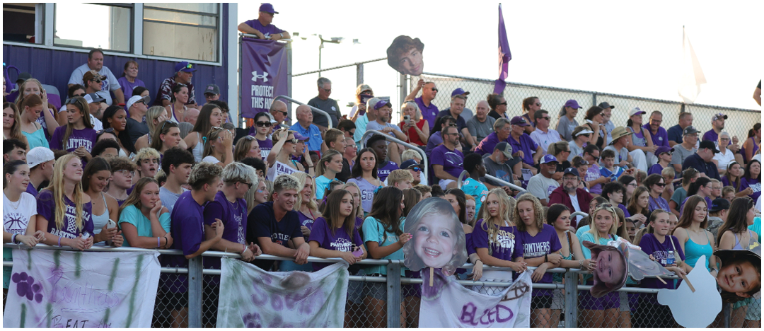 A large crowd of people wearing purple and white clothing is gathered in bleachers at an outdoor event, some holding large cutout faces and banners. Description generated by AI