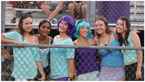 A group of people wearing casual summer clothing stand behind a chain-link fence, some holding phones and keys. Description generated by AI