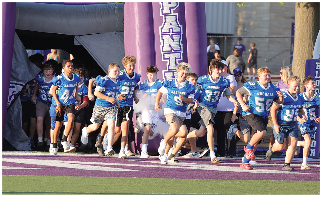 A group of people wearing blue sports jerseys are running out from a large purple inflatable tunnel labeled “PANTHERS" onto a sports field. Description generated by AI