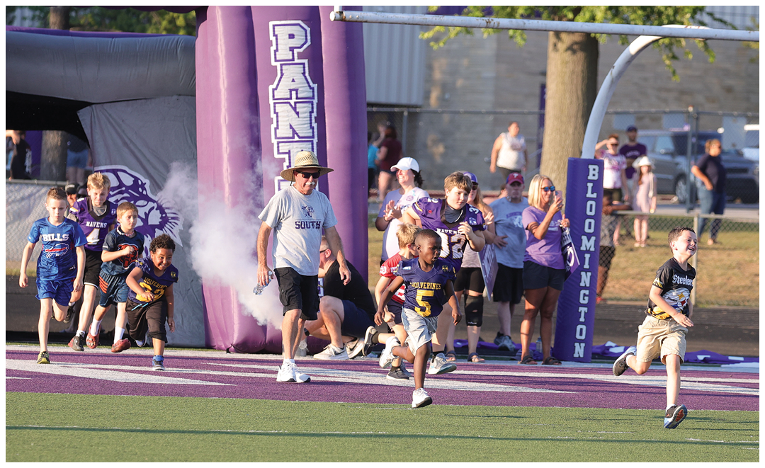 Children in sports uniforms and adults are running and cheering on a football field near an inflatable tunnel and goalpost padding labeled Bloomington. Description generated by AI