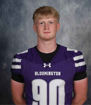 A person with short hair wearing a purple Bloomington sports jersey with the number 90 stands against a gray textured background. Description generated by AI
