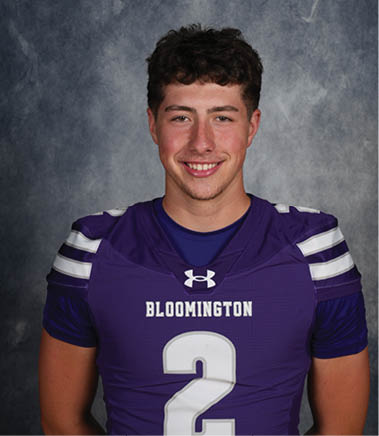 A person with short curly hair is smiling and wearing a purple sports jersey with the word Bloomington and the number 2 on it against a gray textured background. Description generated by AI