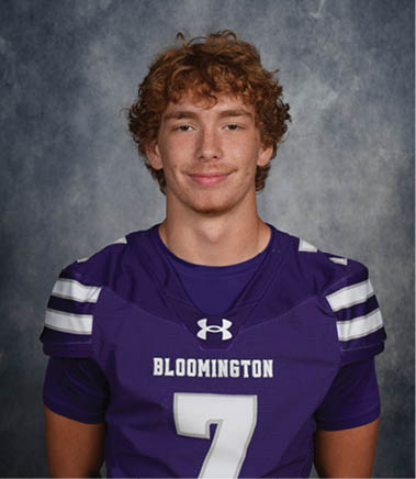 A person with curly hair is wearing a purple sports jersey with the word Bloomington and the number seven on it, standing against a gray textured background. Description generated by AI