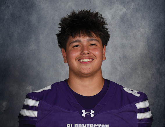 A person with short spiky hair is smiling and wearing a purple sports jersey with the word Bloomington and the number 63 on it against a gray textured background. Description generated by AI