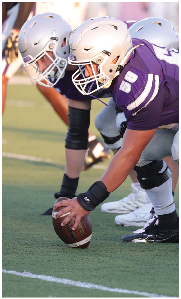 Two people in football helmets and uniforms are positioned on a field with one holding a football ready to snap it. Description generated by AI