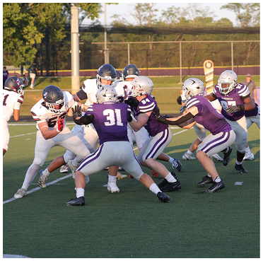 People wearing football uniforms are actively engaged in a game on a green field with trees and a fence in the background. Description generated by AI