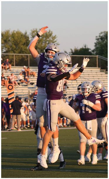Two people in football uniforms jump and celebrate on a field with spectators in the background. Description generated by AI