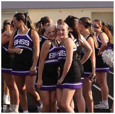 A group of people in matching purple and black cheerleading uniforms with BHSS on the front are standing together outdoors on a sports field, some smiling and posing for the camera. Description generated by AI