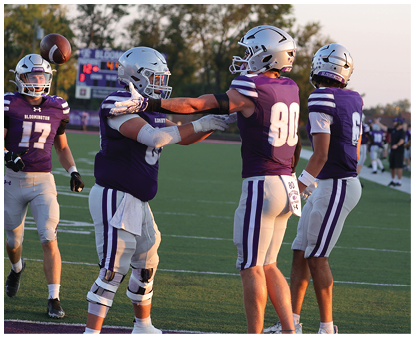 Four people in football uniforms and helmets are on a field with one person throwing a football while the others interact. Description generated by AI