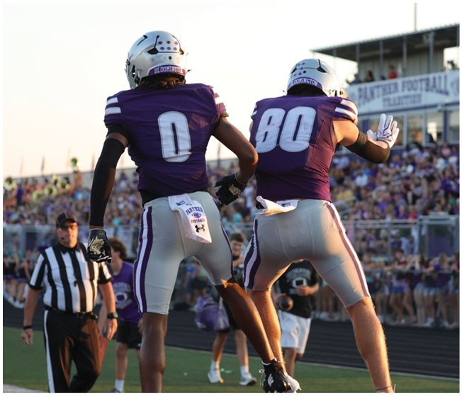 Two football players in purple and white uniforms jump in celebration on a field with a crowd and referee in the background. Description generated by AI