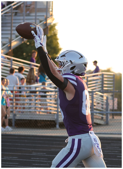 A person wearing a purple and white football uniform and helmet is catching a football with both hands raised above their head on a sports field with bleachers and spectators in the background. Description generated by AI