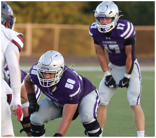 Two people in purple and gray football uniforms and helmets are positioned on a football field, with one crouching and the other standing behind. Description generated by AI