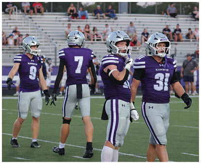 Four people wearing purple and gray football uniforms and helmets stand on a football field with bleachers and spectators in the background. Description generated by AI