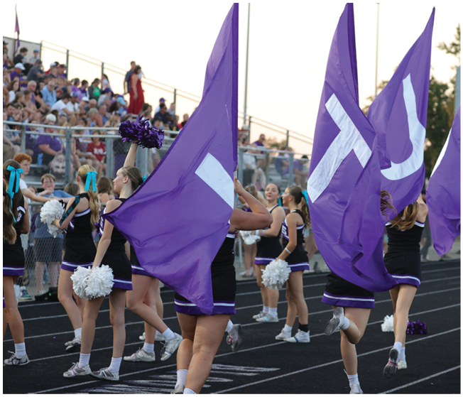 People in cheerleading uniforms are performing with purple flags and pom-poms on a track in front of a crowd seated in bleachers. Description generated by AI