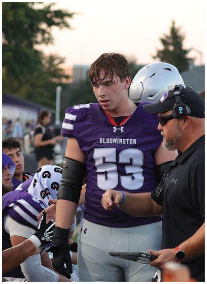 A group of football players in purple uniforms are gathered on the sidelines while a coach wearing a headset and holding a tablet gives instructions. Description generated by AI