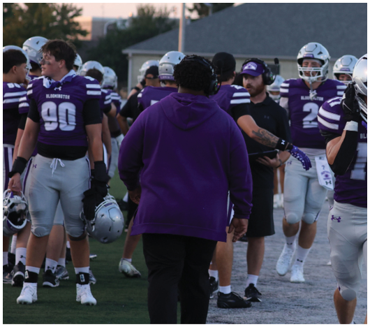 People wearing purple and white football uniforms and helmets are gathered on a field with one person in a purple hoodie walking away from the camera. Description generated by AI