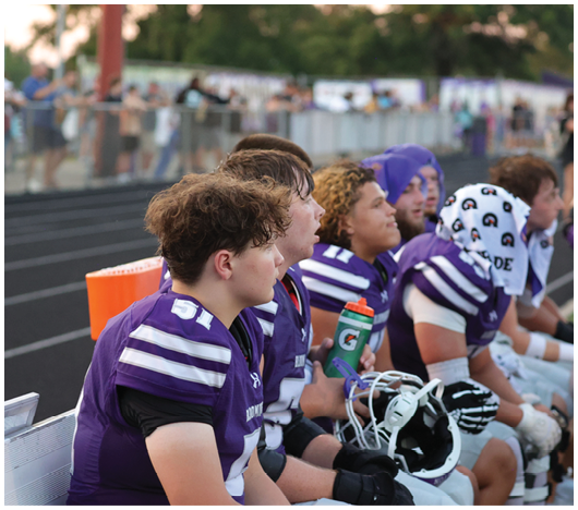 Several people wearing purple and white football uniforms are sitting on a bench at the side of a sports field, some holding helmets and a water bottle. Description generated by AI