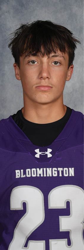 Person with short tousled hair wearing a purple Bloomington sports jersey with the number 23 stands against a gray textured background. Description generated by AI