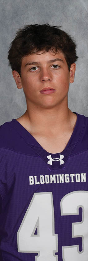 A person with short curly hair wearing a purple Bloomington football jersey with the number 43 stands against a gray textured background. Description generated by AI