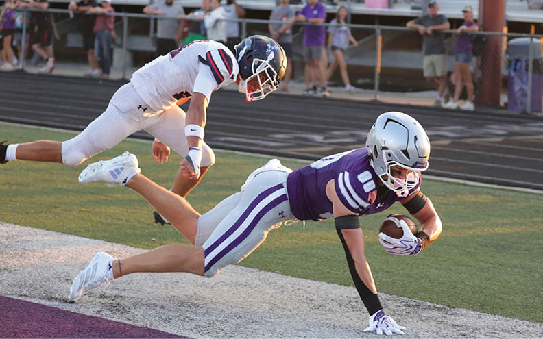 A person in a purple football uniform is diving forward while holding a football as another person in a white uniform reaches to tackle them on a football field. Description generated by AI