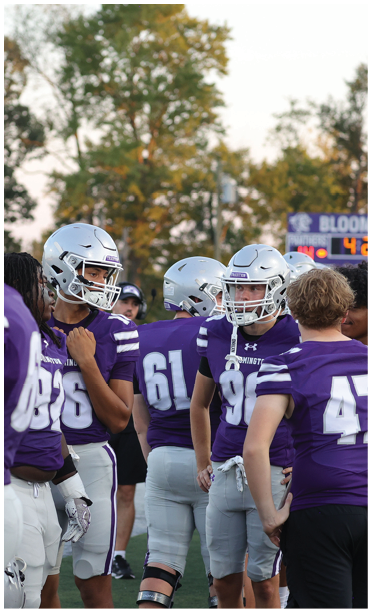 A group of people wearing purple sports jerseys and silver helmets are standing together on a field with trees and a scoreboard in the background. Description generated by AI