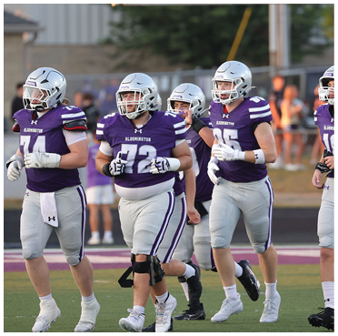 Four people wearing purple and white football uniforms and helmets are walking on a field with a blurred background of other people and structures. Description generated by AI