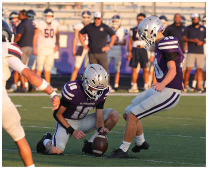 Two people in football uniforms and helmets are on a field with one holding a football upright while the other prepares to kick it. Description generated by AI
