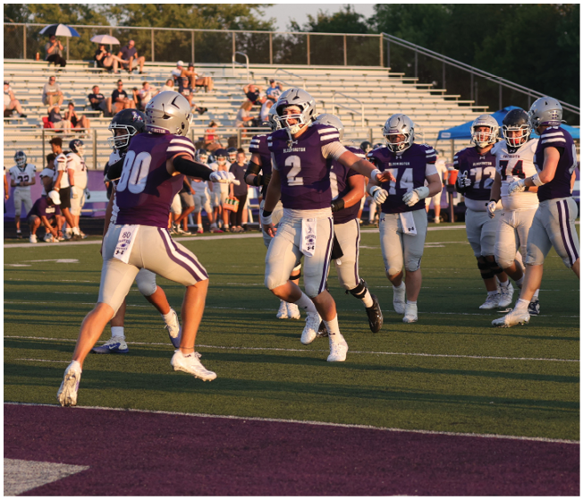 People wearing purple and white football uniforms are running and celebrating on a football field with empty bleachers in the background. Description generated by AI