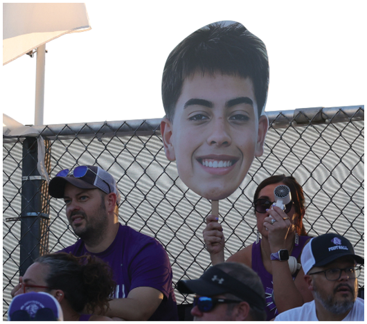 Two adults stand behind a chain-link fence holding a large cutout of a smiling person's face on a stick. Description generated by AI