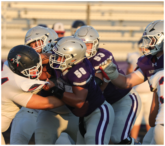 Several people wearing football helmets and uniforms are engaged in a close physical play on a football field with empty bleachers in the background. Description generated by AI