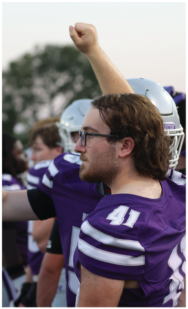 People wearing purple football uniforms stand on the sidelines with one person raising a fist in the air. Description generated by AI