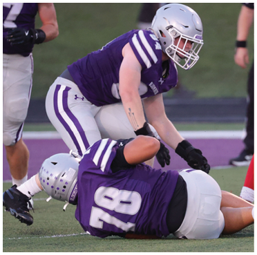 Two people in purple and white football uniforms are on a field with one person lying on the ground and the other leaning over them. Description generated by AI