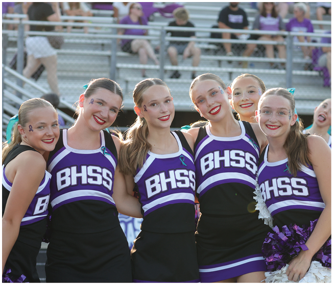 Five people wearing matching purple and black cheerleading uniforms with BHSS on the front are standing close together smiling in an outdoor stadium setting. Description generated by AI
