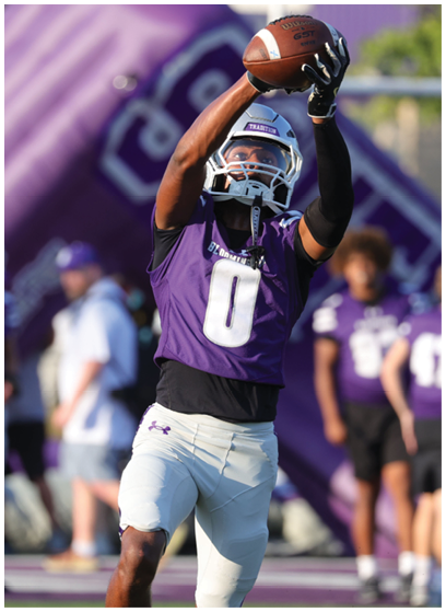 A person wearing a purple sports jersey and white football pants is running on a field with other people in sports attire blurred in the background. Description generated by AI
