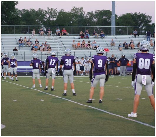 Several people in purple and white football uniforms stand spaced apart on a green field with a crowd and other players in the background. Description generated by AI