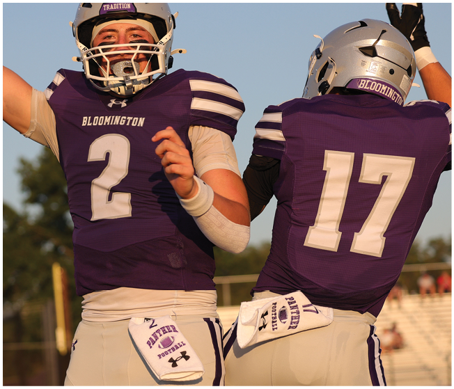 Two football players in purple Bloomington jerseys celebrate on the field during a game. Description generated by AI