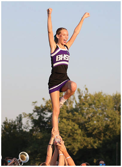A person wearing a BHSS cheerleading uniform with a ribbon face sticker is raising both arms enthusiastically against a clear sky. Description generated by AI