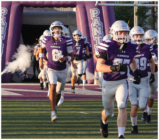 Several people wearing football helmets are standing under a large purple inflatable arch with white and purple lettering. Description generated by AI