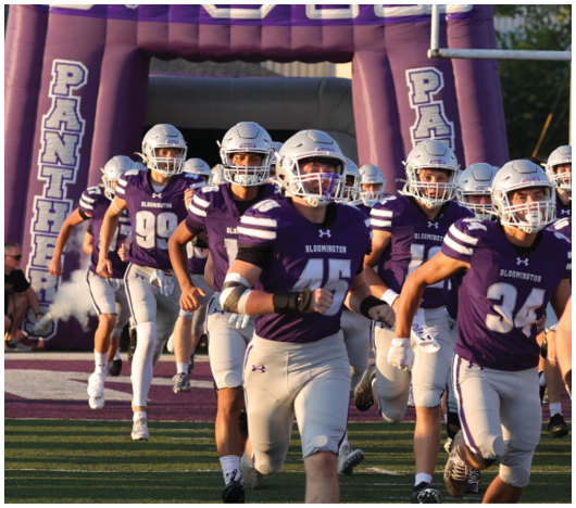 A large purple inflatable arch with the word “UTCH" and a bear logo stands outdoors near a football goalpost with people wearing helmets partially visible below. Description generated by AI