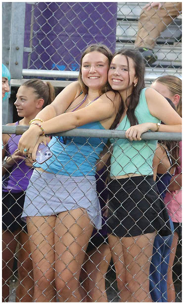 Two people smiling and hugging each other while leaning on a metal fence with other people and bleachers in the background. Description generated by AI
